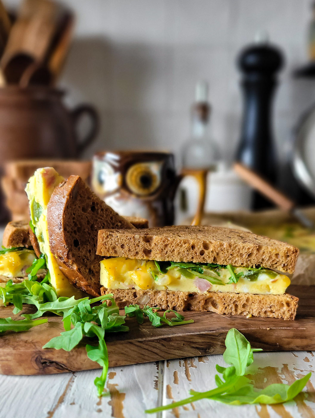 A toasted Western Sandwich is made and sitting on a cutting board, with arugula strewn around, and a mug of coffee in the background.