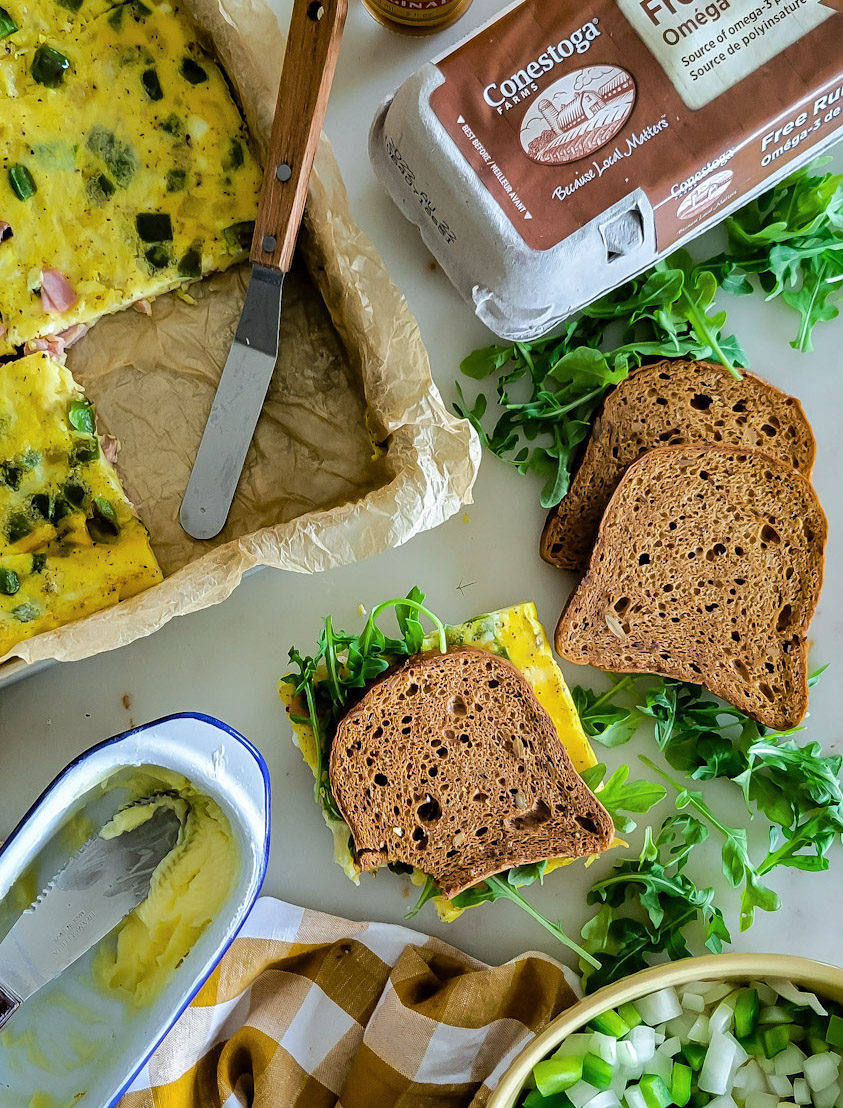 Toasted Western Sheet Pan Baked Eggs, with finished sandwiches and a carton of Conestoga Eggs in the corner. Arugula is strewn about, and butter and a knife are in the bottom corner.