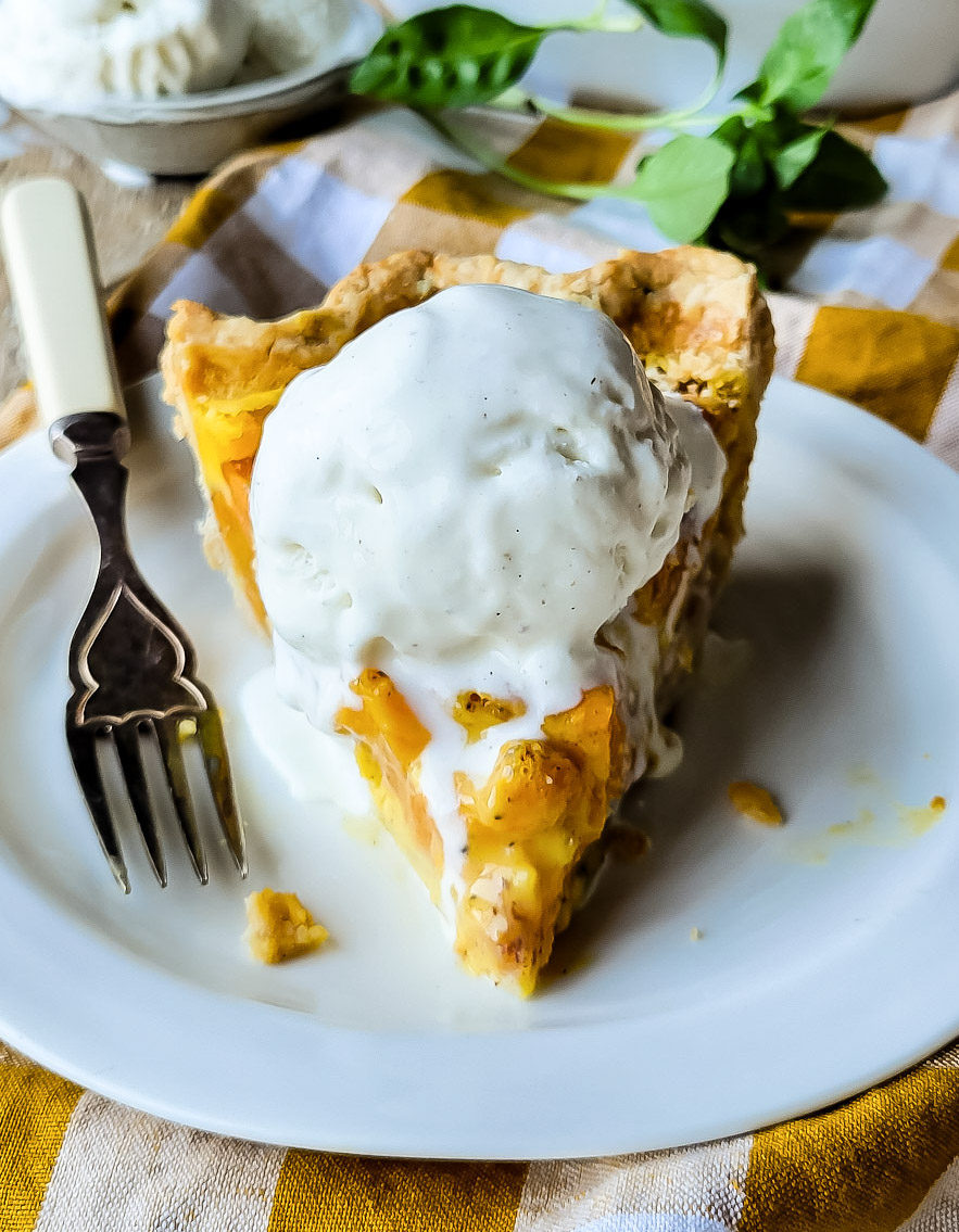 Close up of a slice of peach sour cream pie on a plate with vanilla ice cream, basil leaves in the background.