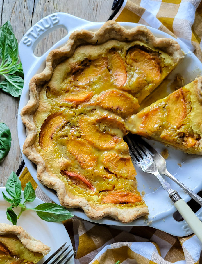 Peach Sour Cream Pie in a Basil Crust, a slice on a plate next to the pie with basil leaves surrounding everything.