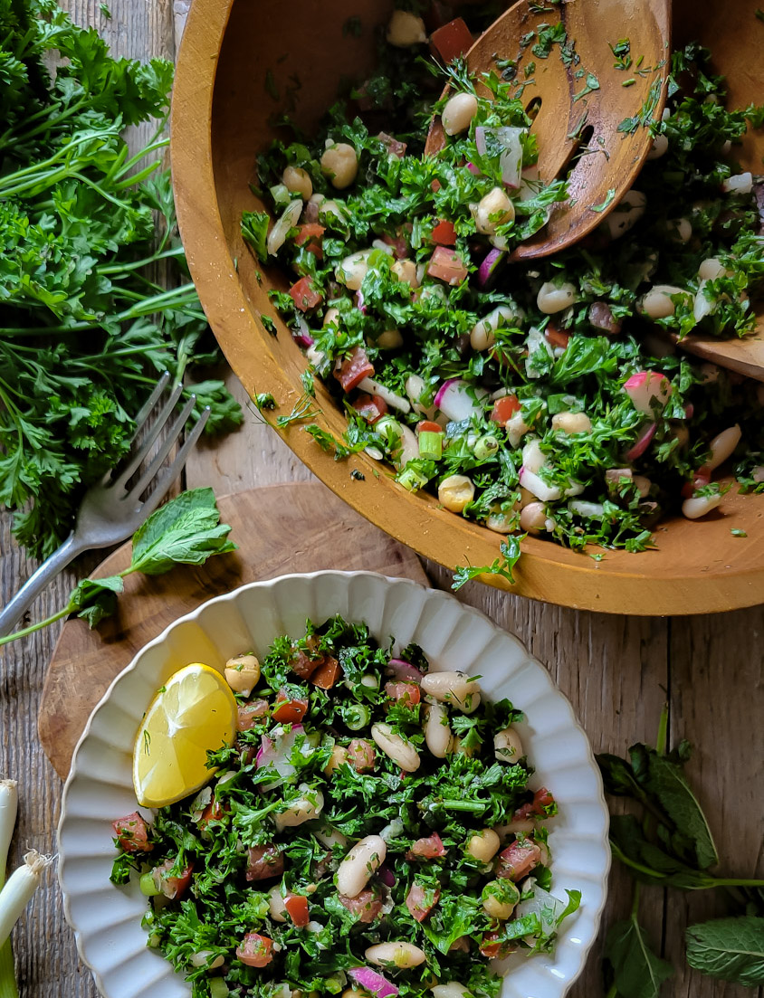 Wooden Bowl of parsley and mint white bean tabouleh salad with a plate of the salad next to it. Parsley and mint leaves are on the side.