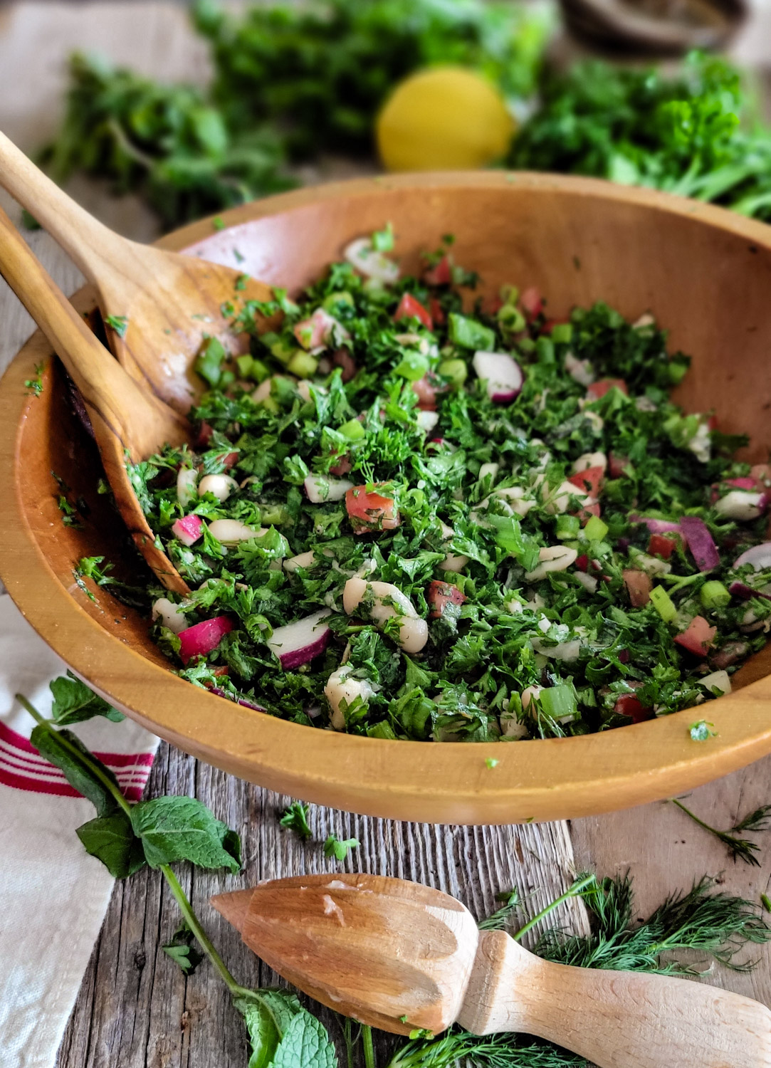 Wooden Bowl of parsley and mint white bean tabouleh salad with a plate of the salad next to it. Parsley and mint leaves are on the side.