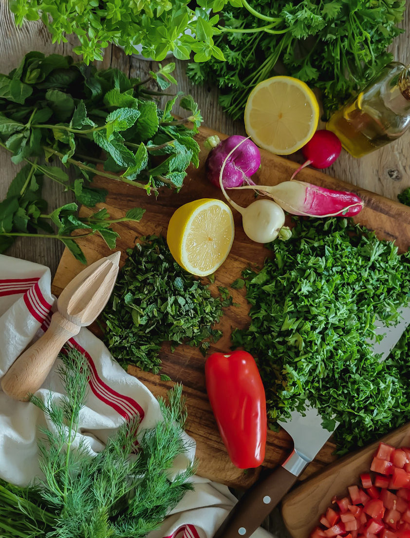 Cutting board with fresh parsley, mint, tomatoes, radishes and tomatoes scattered on top.