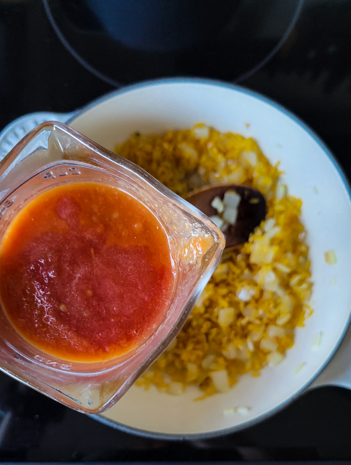 A glass measuring cup with tomato puree about to be poured into a pot with sautéed onion, garlic and toasted orzotto.