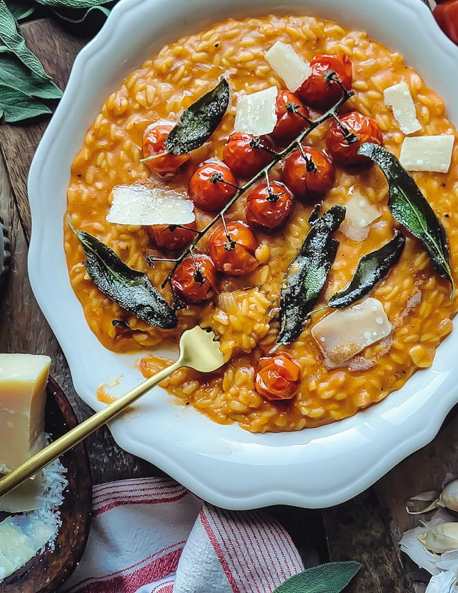 A serving plate filled with creamy tomato orzotto with roasted tomatoes on top, sage leaves and parmesan cheese on the side.