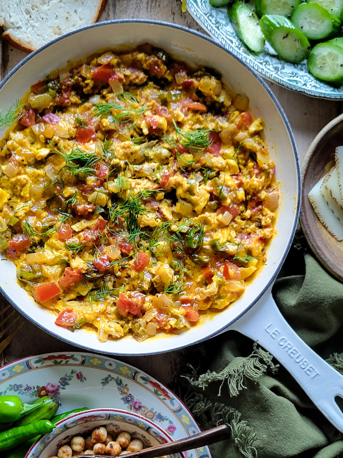 A skillet with turkish scrambled eggs, made of eggs, tomatoes, shishito peppers and onions. Sliced cucumbers and peppers are on the side.