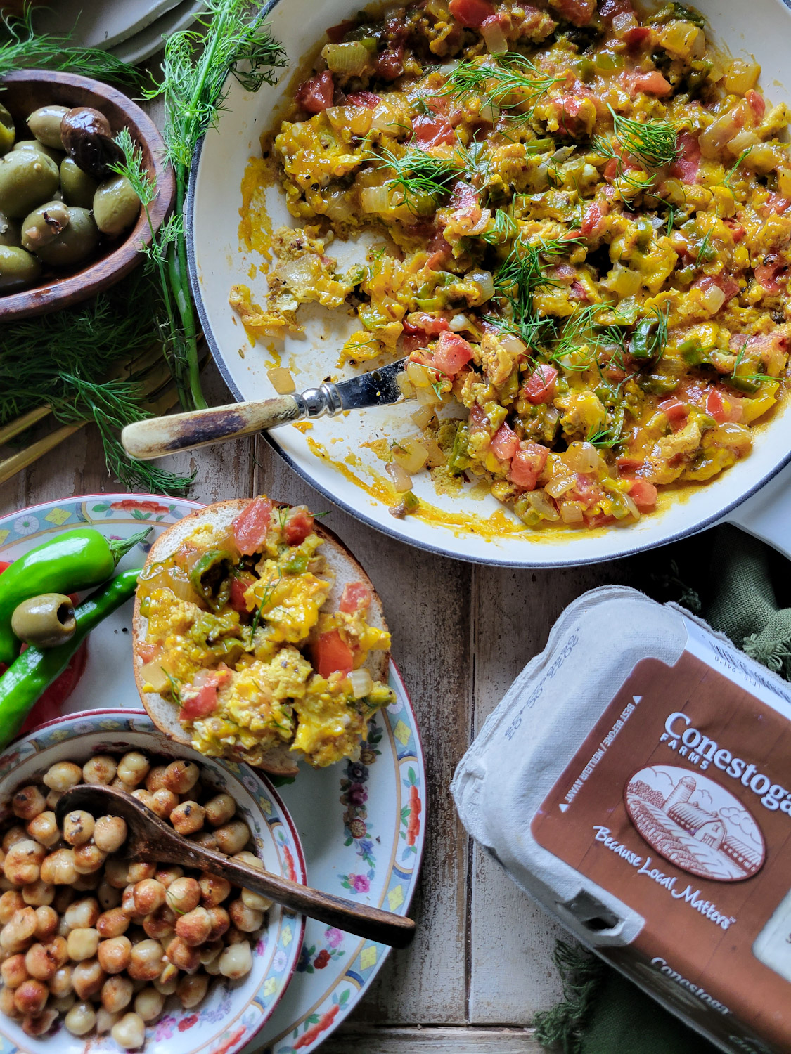 A skillet with Turkish scrambled eggs, some spread on a bagel, with olives and spiced chickpeas on the side.