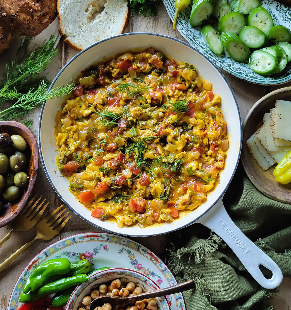 A Levantine breakfast table set with a skillet of Turkish Scrambled Eggs, slice cucumbers, bagels, cheeses, olives and spiced chickpeas surrounding it.