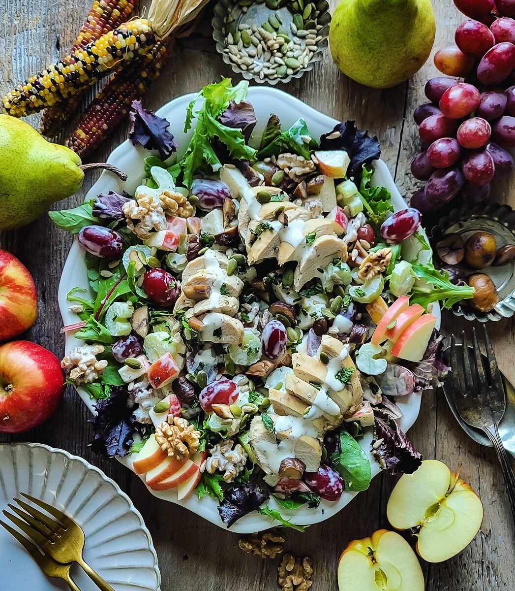 A platter filled with a crunchy colorful Waldorf Salad, with apples, grapes, celery, chicken and chestnuts. The platter is surrounded by rainbow corn, apples, pumpkin and sunflower seeds and grapes.