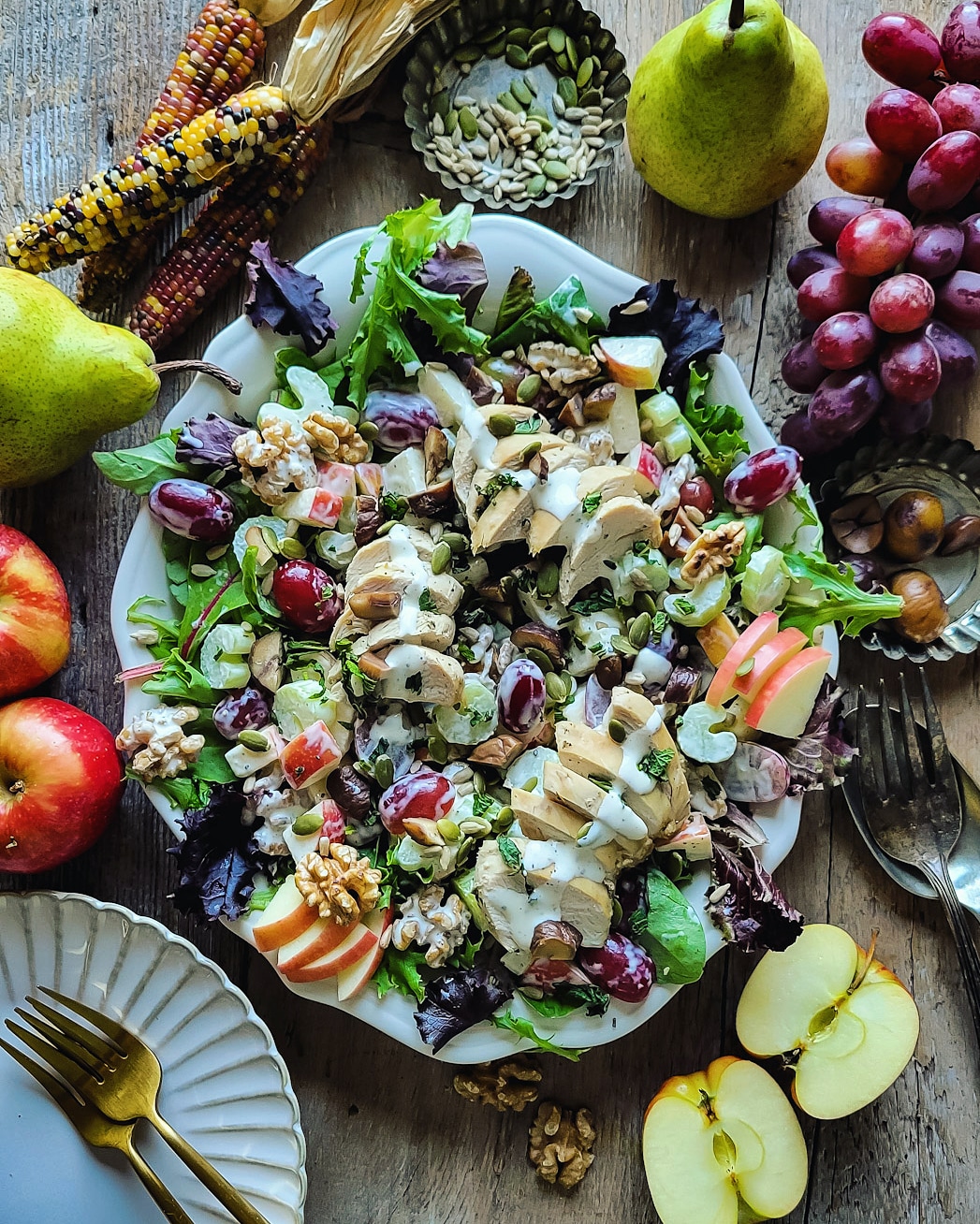 A platter filled with a crunchy colorful Waldorf Salad, with apples, grapes, celery, chicken and chestnuts. The platter is surrounded by rainbow corn, apples, pumpkin and sunflower seeds and grapes.