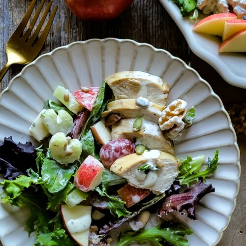Close up of a plate filled with crunchy, colorful Waldorf Salad, with apples, grapes, celery, chicken and chestnuts. Off in a corner is an apple and fork, as well as a platter with more salad.