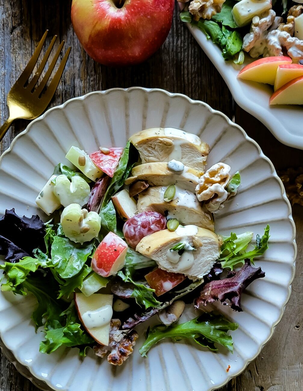 Close up of a plate filled with crunchy, colorful Waldorf Salad, with apples, grapes, celery, chicken and chestnuts. Off in a corner is an apple and fork, as well as a platter with more salad.