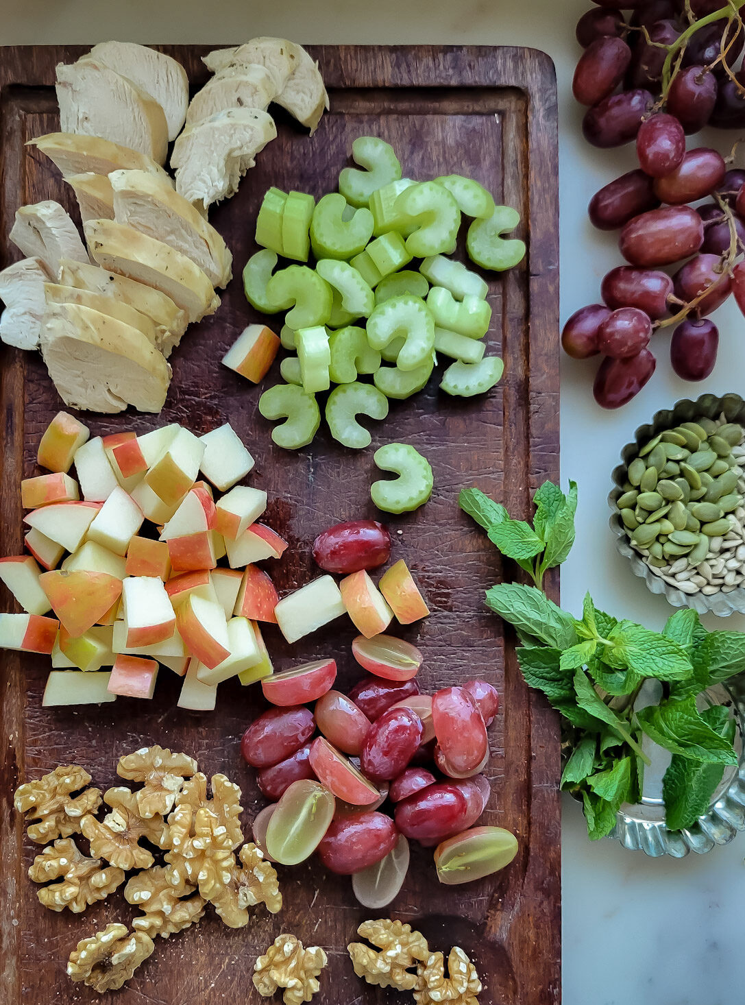 Ingredients for Waldorf Salad on a cutting board. Poached chicken; diced celery, apples and grapes as well as walnuts, mint, and pumpking seeds.