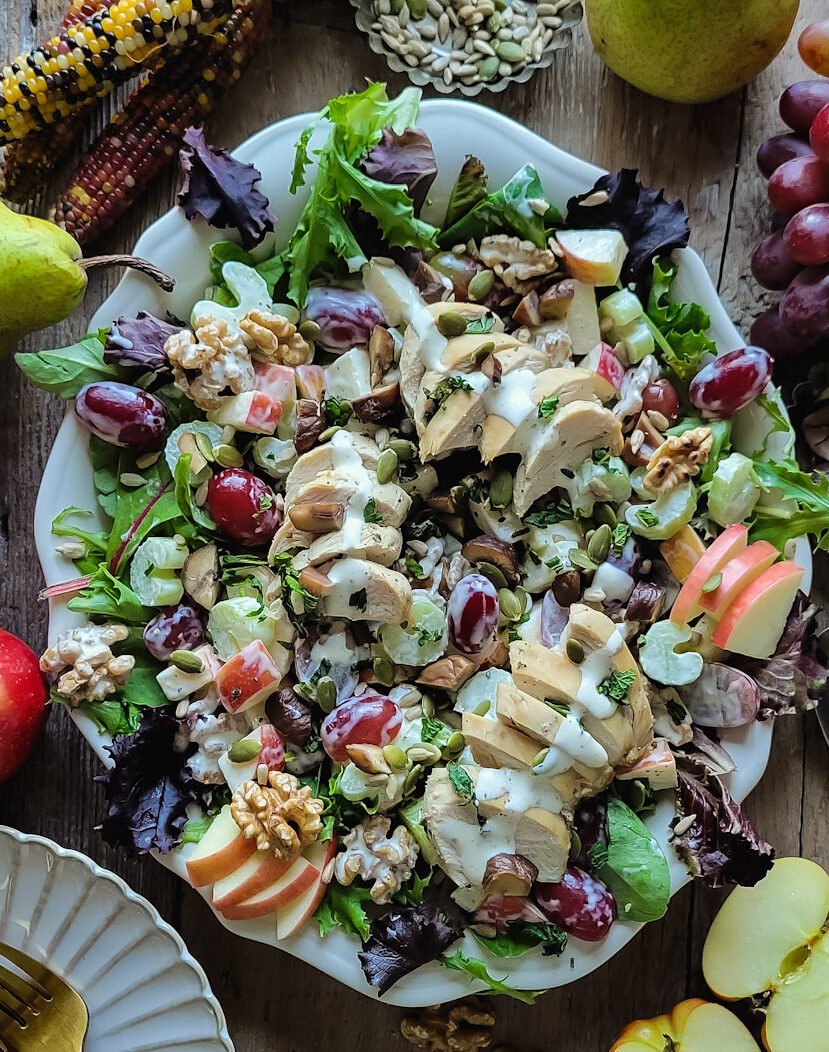 A platter filled with a crunchy colorful Waldorf Salad, with apples, grapes, celery, chicken and chestnuts. The platter is surrounded by rainbow corn, apples, pumpkin and sunflower seeds and grapes.