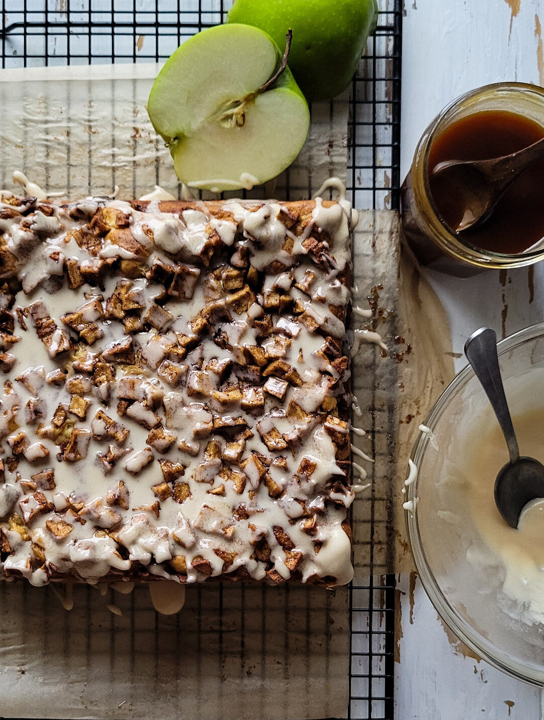 Cooling rack with apple fritter squares on it. Salted bourbon caramel sauce and glaze are to the side, and a granny smith apple is sitting on the cooling rack as well.