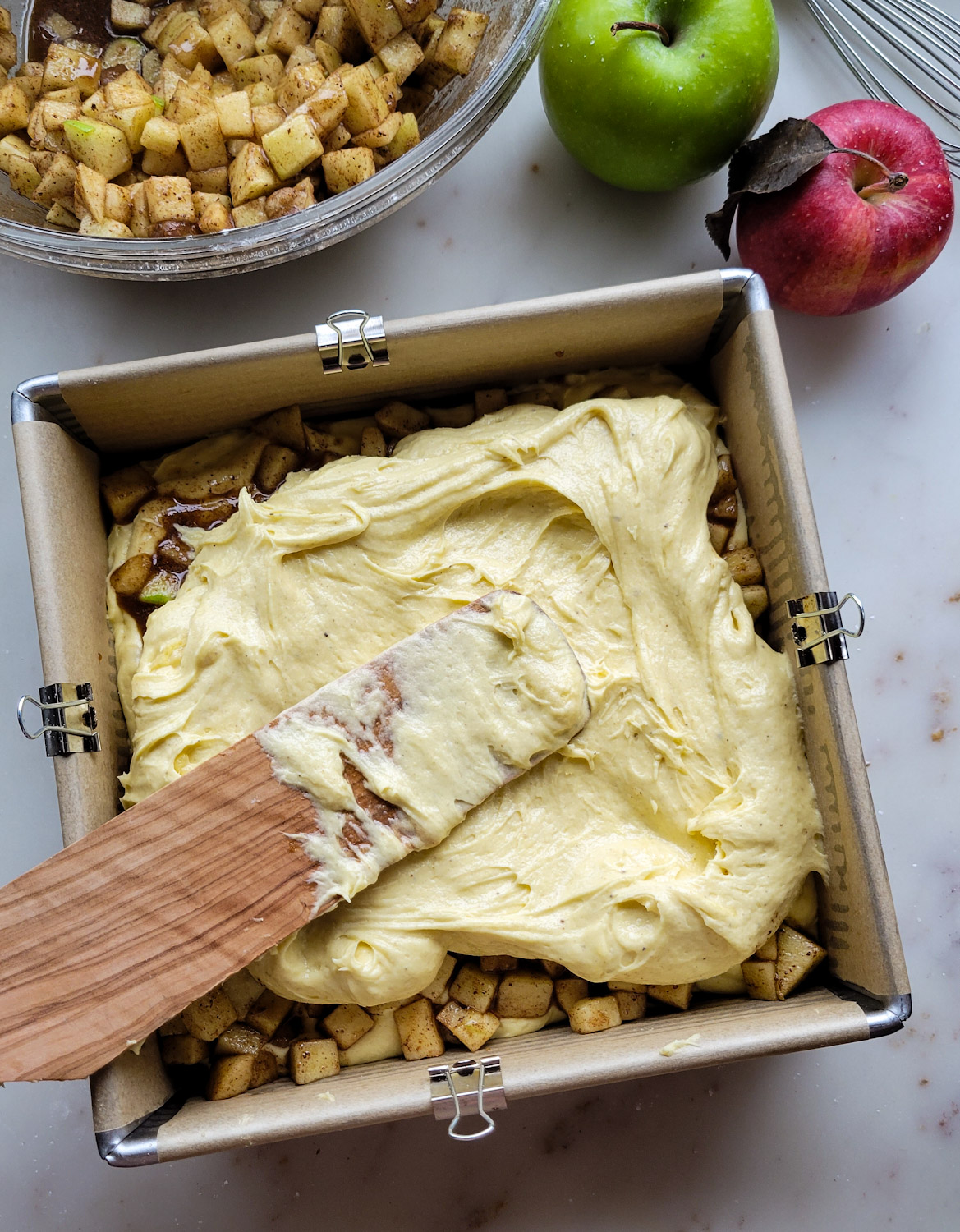 A baking pan with layers of cake batter and diced spiced apples. A bowl of diced spiced apples is to the side, as well as two fresh apples.
