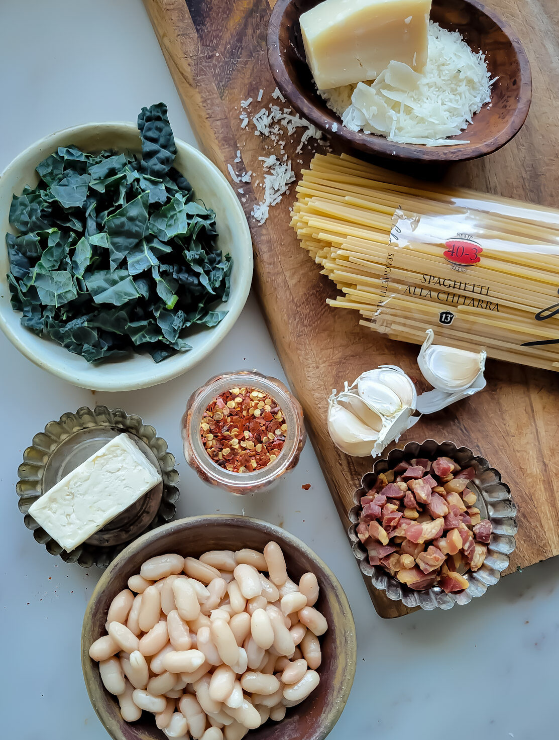 Ingredients needed to make Brown Butter Bean and Kale Pasta: a bowl of chopped Kale, grated parmesan cheese, spaghetti, crispy pancetta pieces, garlic, chilli flakes, butter and Cannellini beans