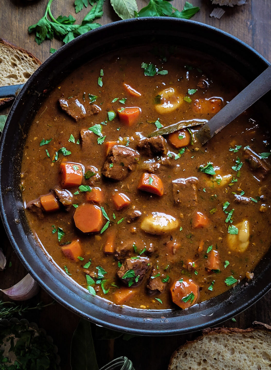 A pot filled with rich and hearty Goulash Soup, filled with beef, carrots, gnocchi, thyme and parsley. There are slices of sourdough bread, and fresh parsley scattered around the pot.