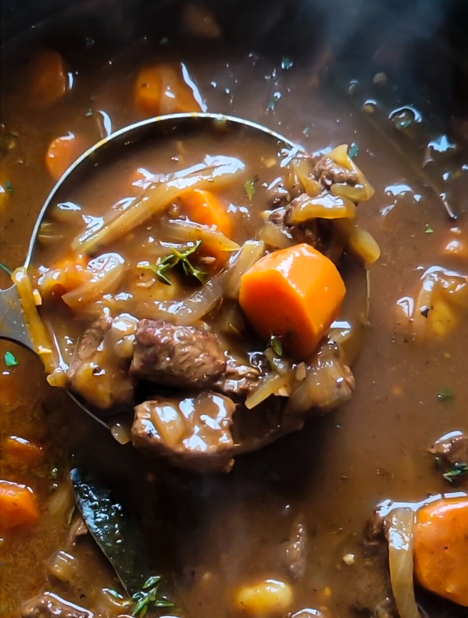 Close up of a ladle holding Goulash Soup, or Gulaschsuppe. The ladle is filled with beef, onions, carrots, thyme and broth.