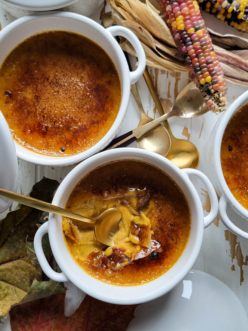 Three pots of pumpkin creme brùlée with autumn leaves and rainbow corn nearby. A spoon has cracked through the sugar shell to expose the custard beneath.