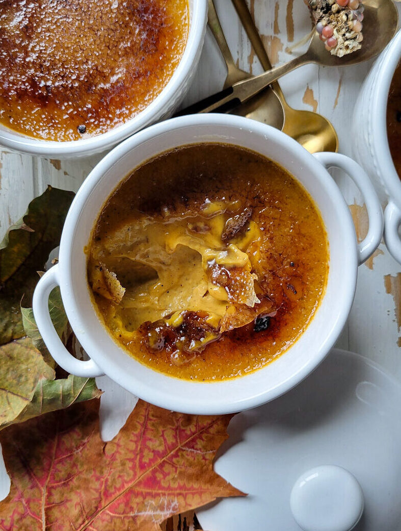 Three pots of pumpkin creme brùlée with autumn leaves and rainbow corn nearby. A spoon has cracked through the sugar shell to expose the custard beneath.