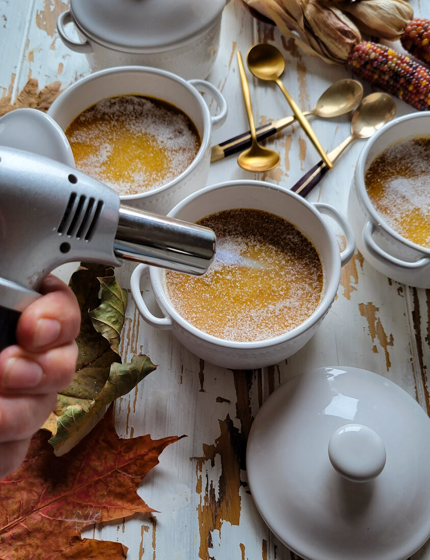 three small pots of pumpkin creme brûlée on a table, with a torch aimed at one of them.