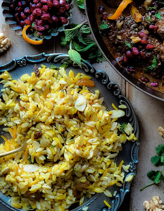 A plate of jewelled saffron Orzo with Chicken Fessenjan, Persian Walnut and Pomegranate Stew. Pomegranate arils and mint leaves are scattered around the dish.