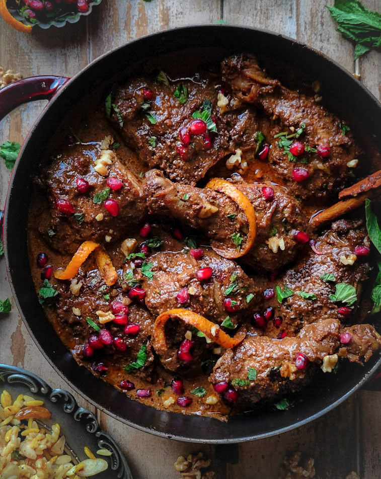 A plate of jewelled saffron Orzo with Chicken Fessenjan, Persian Walnut and Pomegranate Stew. Pomegranate arils and mint leaves are scattered around the dish.