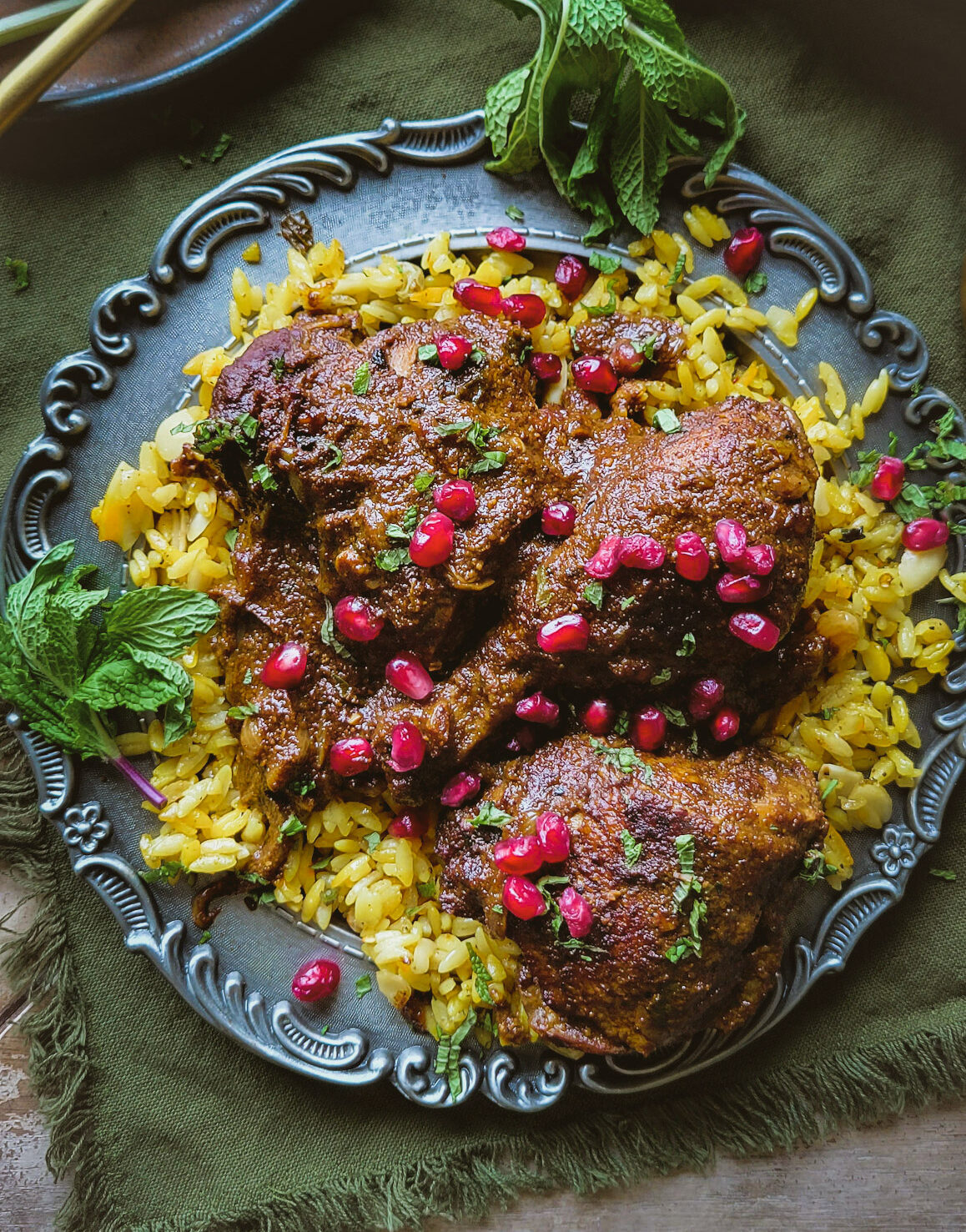 A plate of jewelled saffron Orzo with Chicken Fessenjan, Persian Walnut and Pomegranate Stew. Pomegranate arils and mint leaves are scattered around the dish.