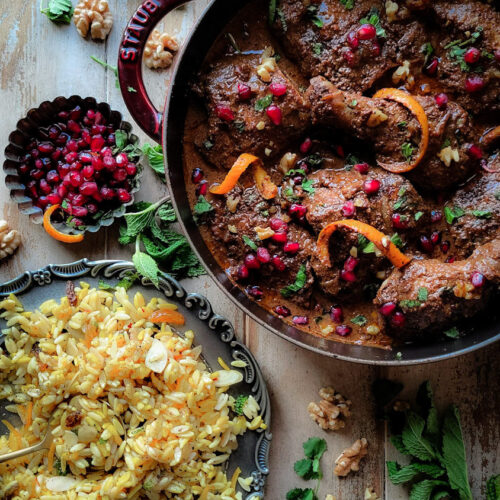 A plate of jewelled saffron Orzo with Chicken Fessenjan, Persian Walnut and Pomegranate Stew. Pomegranate arils and mint leaves are scattered around the dish.