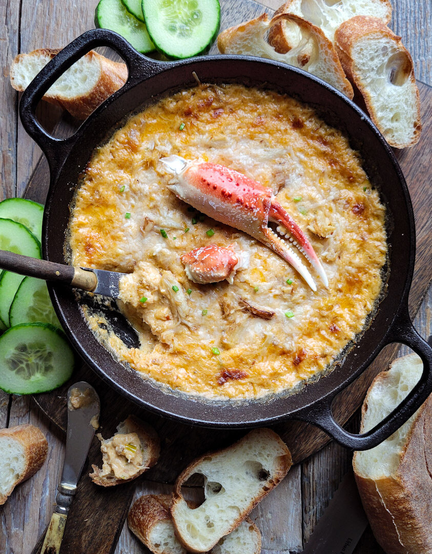 A cast iron skillet filled with Harissa Crab and Cheddar Dip, garnished with a crab claw. Sliced baguette and cucumbers surround the dip.