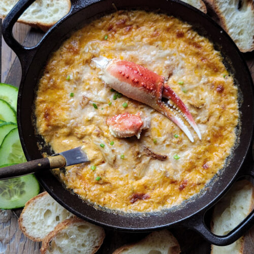 A cast iron skillet filled with Harissa Crab and Cheddar Dip, garnished with a crab claw. Sliced baguette and cucumbers surround the dip.