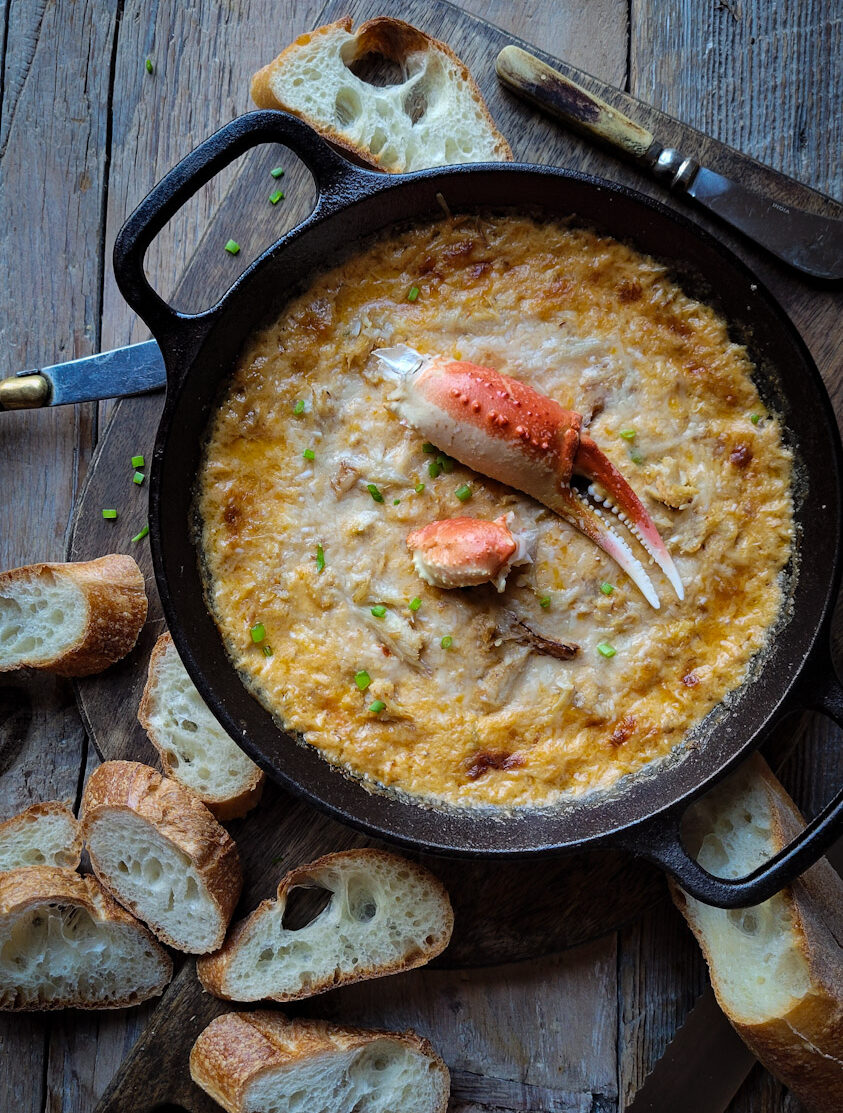 A cast iron skillet filled with Harissa Crab and Cheddar Dip, garnished with a crab claw. Sliced baguette surrounds the dip.