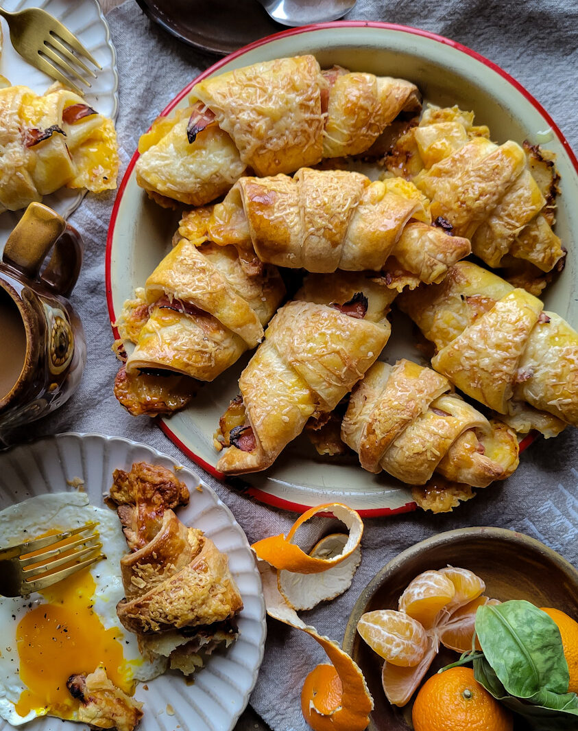 A platter of ham and cheese croissants on a breakfast table. Coffee, orange juice, clementines and plates with eggs are on the table as well.