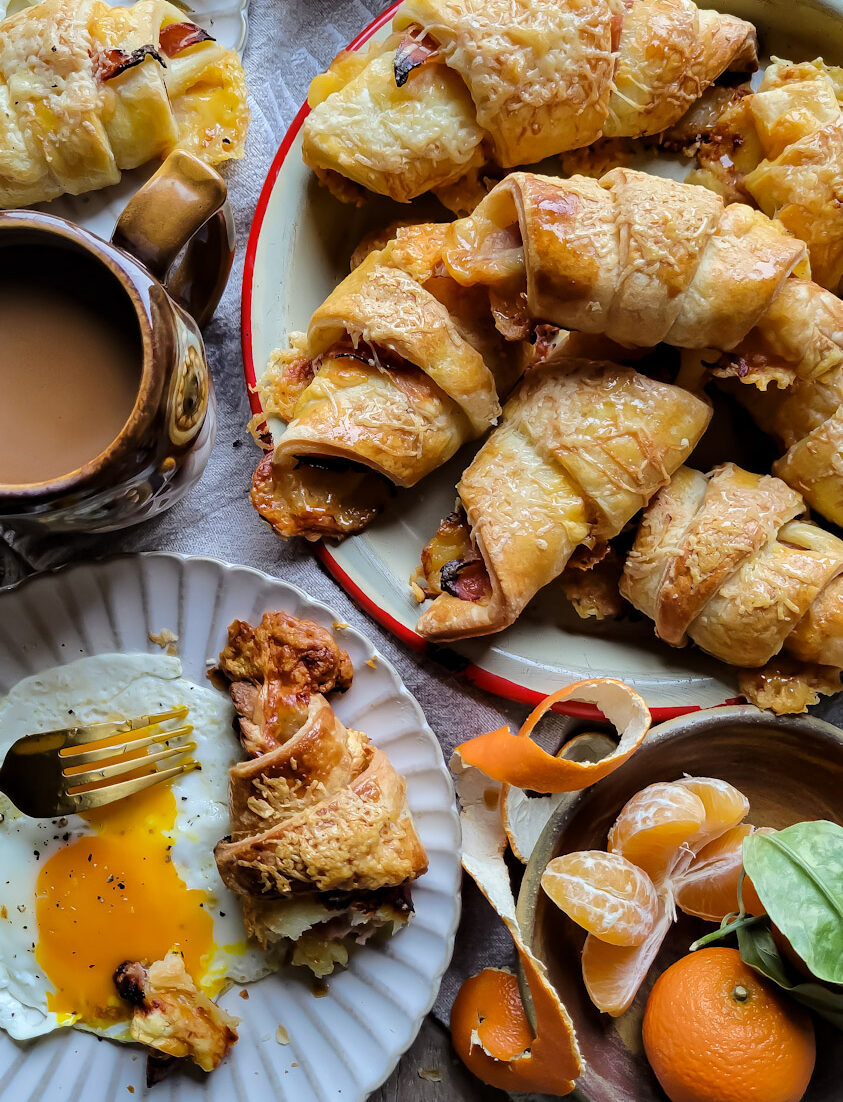 A platter of ham and cheese croissants on a breakfast table. Coffee, orange juice, clementines and plates with eggs are on the table as well.