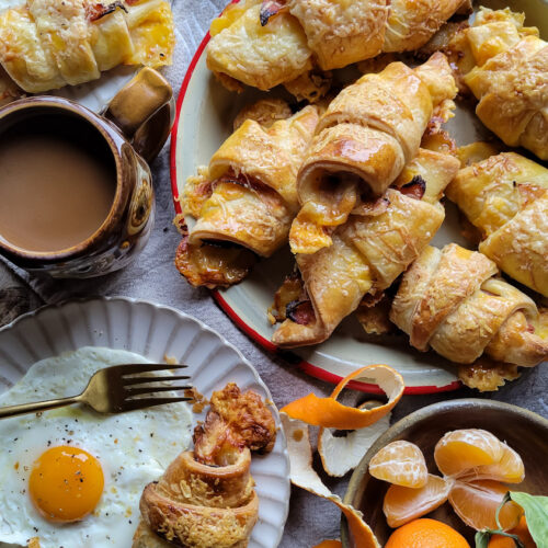 A platter of ham and cheese croissants on a breakfast table. Coffee, orange juice, clementines and plates with eggs are on the table as well.