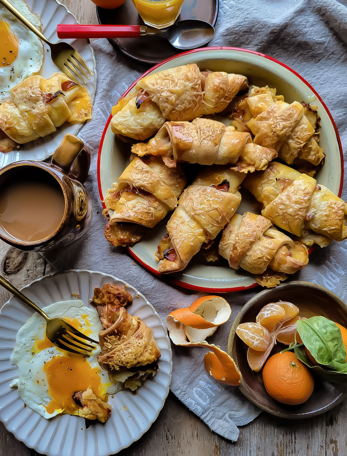 A platter of ham and cheese croissants on a breakfast table with plates of eggs, clementines and coffee cup surrounding it.