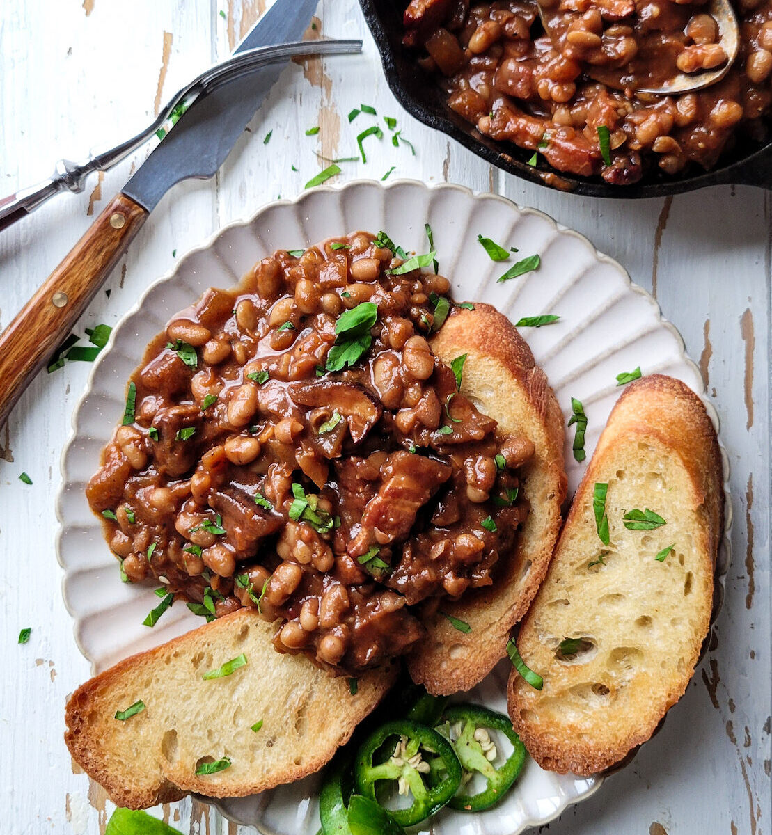 a plate with Classic Baked Beans on Toast, with the skillet of baked beans to the side. The plate is garnished with parsley and peppers.