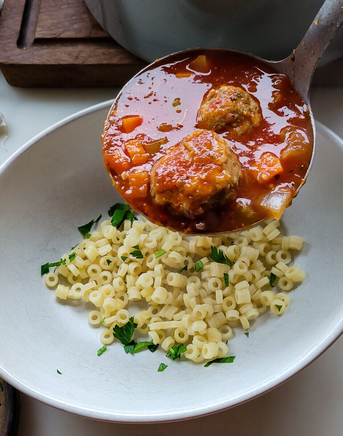 A ladle with Meatball Soup being poured over a bowl with pasta in it.