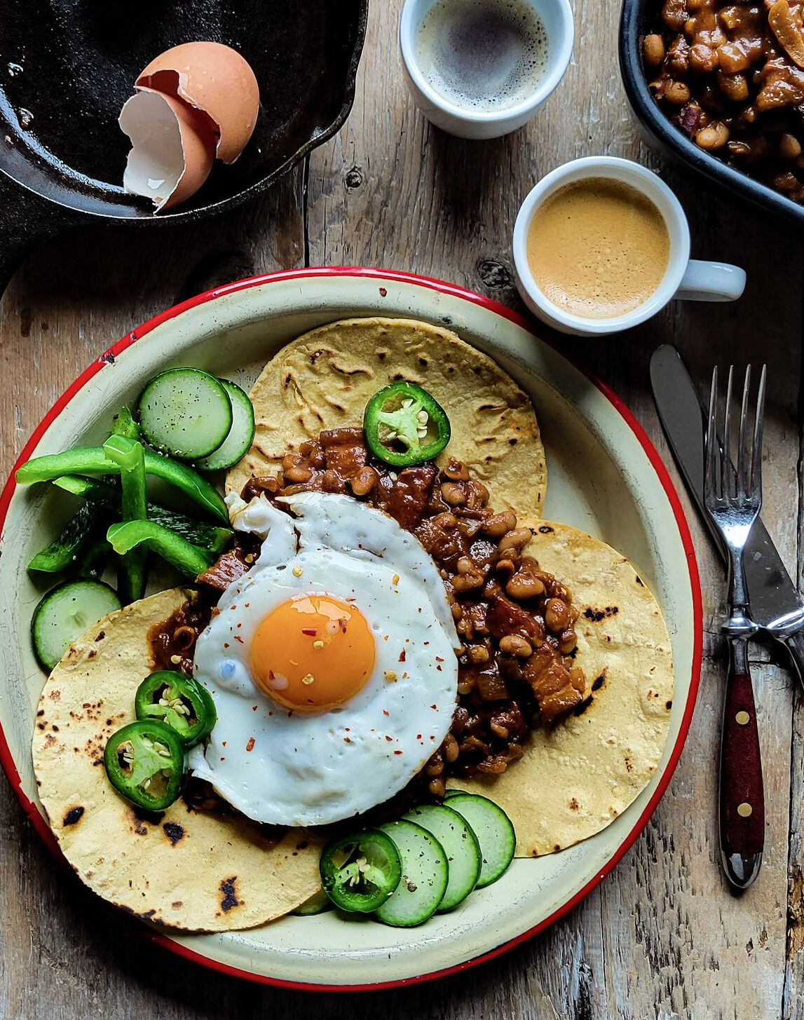 A plate with tortillas, baked beans,and an egg for breakfast, with coffee, a skillet with egg shells and more baked beans are to the side.