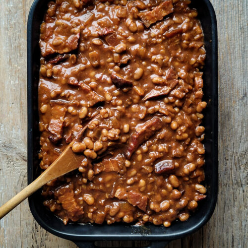 A casserole baking dish filled with Classic Baked Beans on a wood surface, with a wooden spoon is to the side.