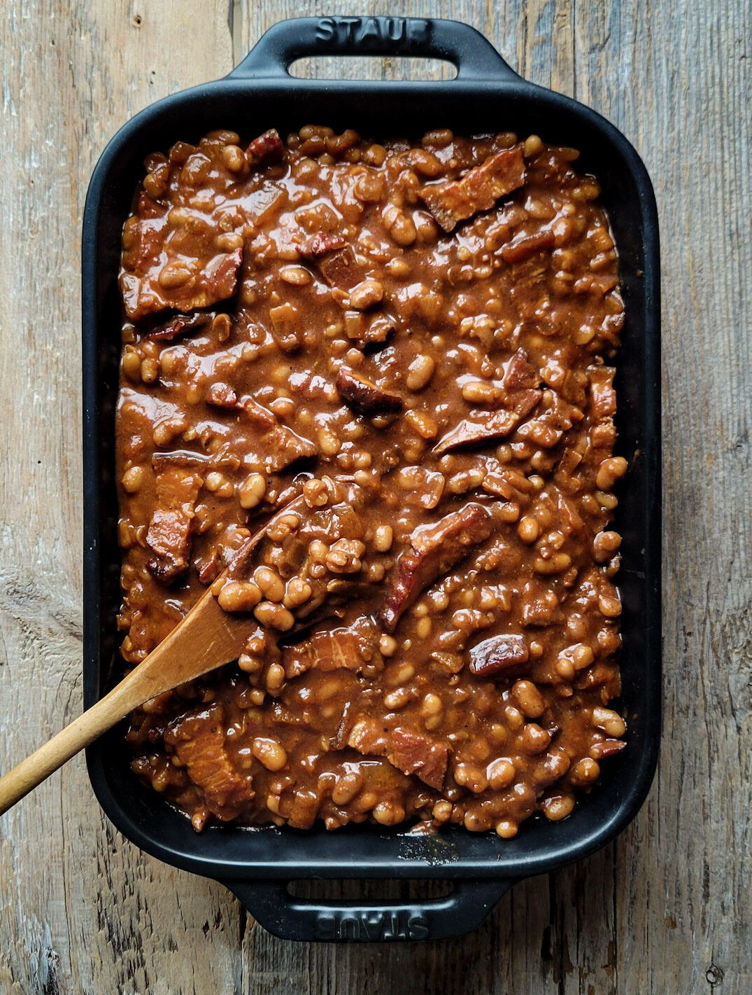 A casserole baking dish filled with Classic Baked Beans on a wood surface, with a wooden spoon is to the side.
