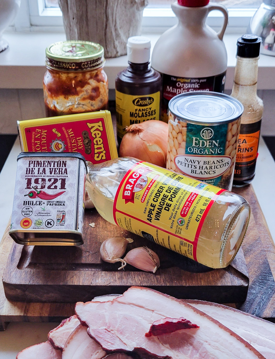 Ingredients for Classic Baked Beans on a cutting board: BBQ sauce, molasses, maple syrup, Worcestershire sauce, can of beans, onion, dry mustard, smoked paprika, apple cider vinegar, garlic and thick cut bacon.