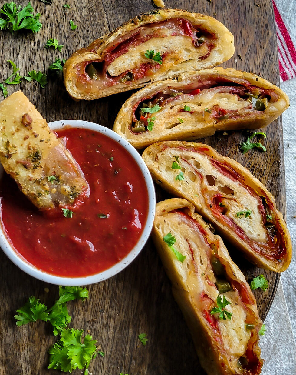 A cutting board with sliced stromboli (rolled pizza) with harissa dipping sauce, one slice dipped into the bowl.