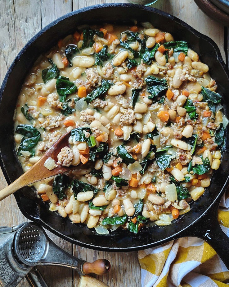 A cast iron skillet filled with Brothy Beans with Sausage and Kale, a cheese grater to the side.