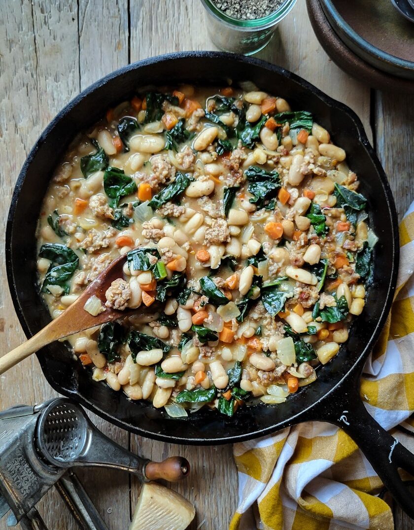 A skillet filled with Brothy Beans with Sausage and Kale, a cheese grater to the side.