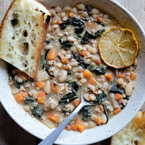 A bowl containing a generous portion of Brothy Beans with Sausage and Kale, with a slice of baguette and charred lemon slice on the side.
