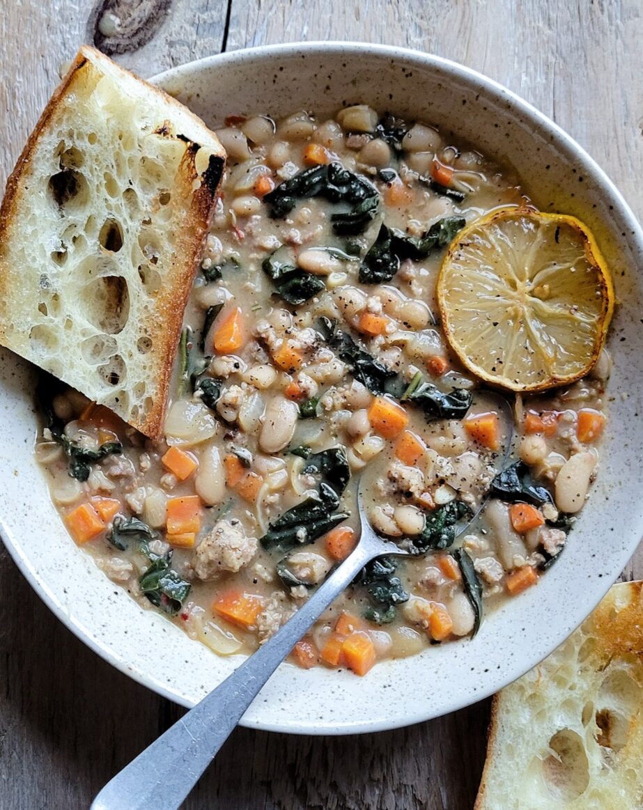 A bowl containing a generous portion of Brothy Beans with Sausage and Kale, with a slice of baguette and charred lemon slice on the side.