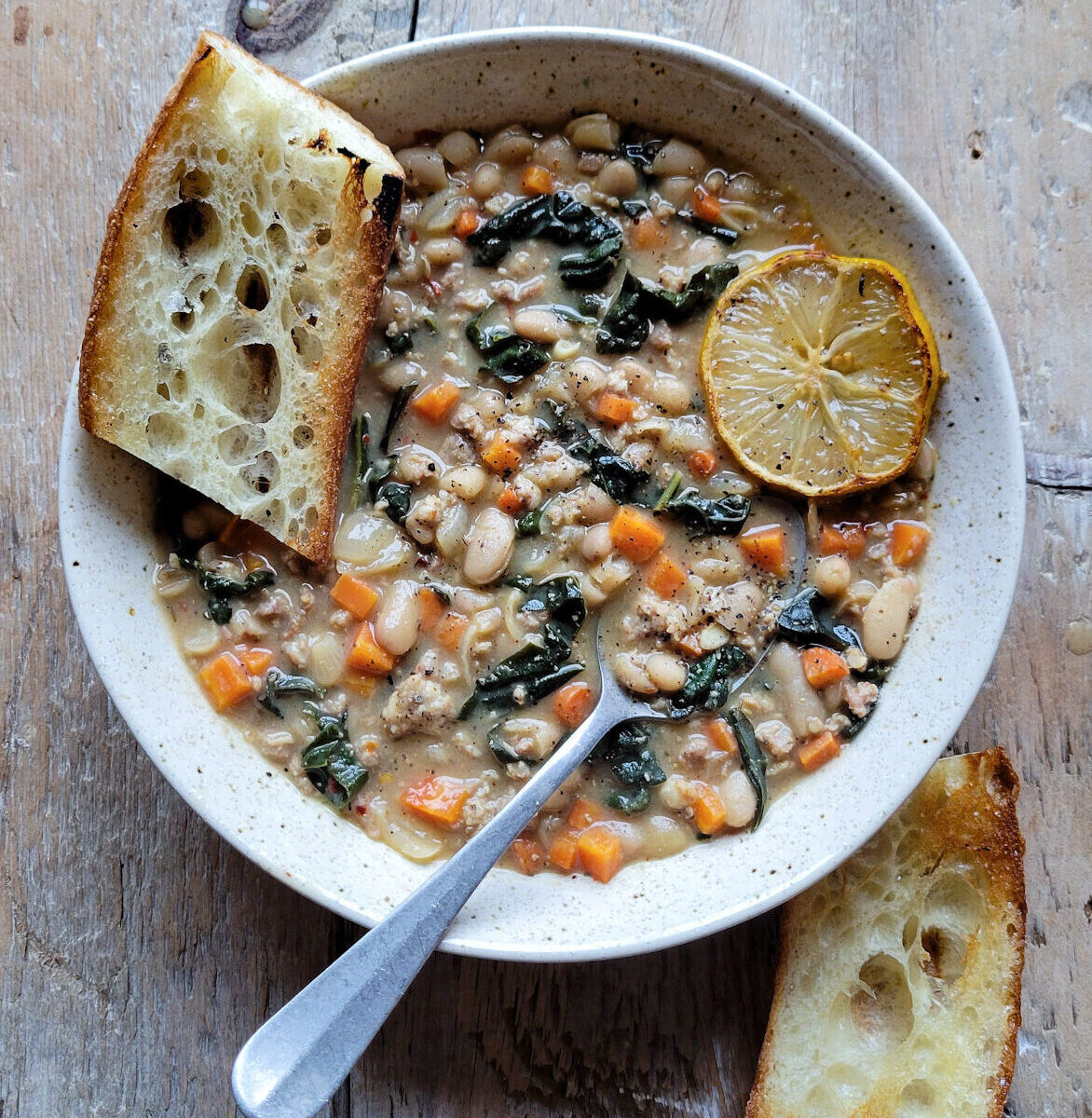 A bowl of Brothy Beans with Sausage and kale on the table, with toated baguette slices and lemon on the side.