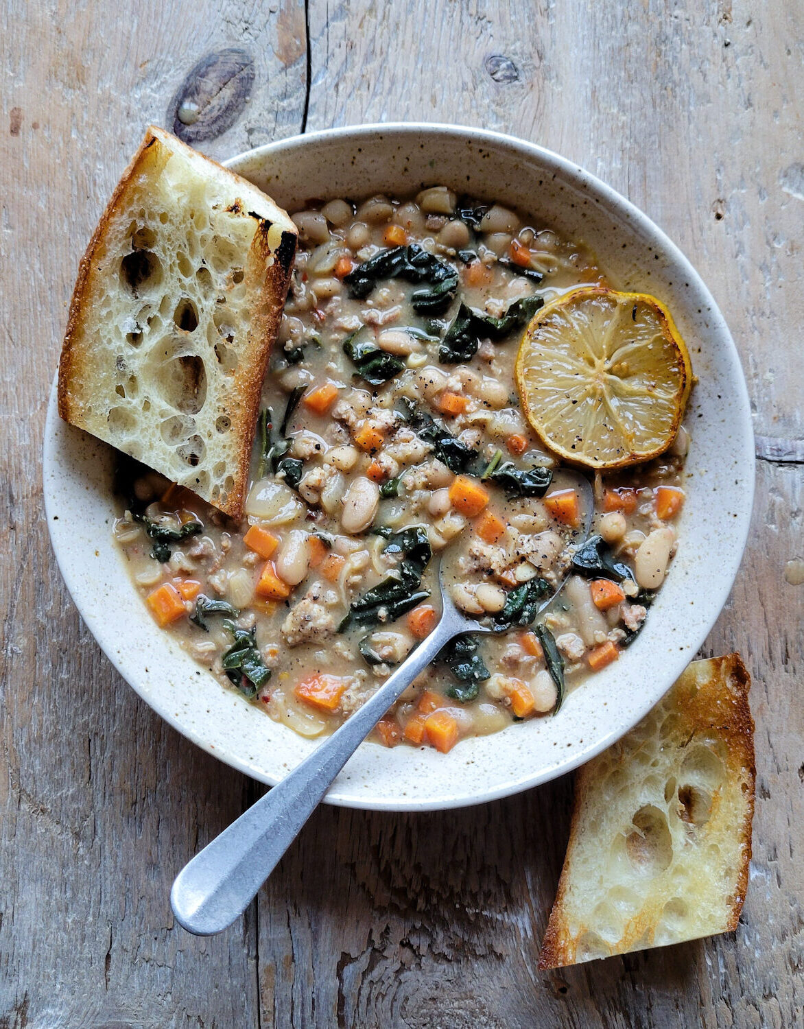 A bowl of Brothy Beans with Sausage and kale on the table, with toated baguette slices and lemon on the side.